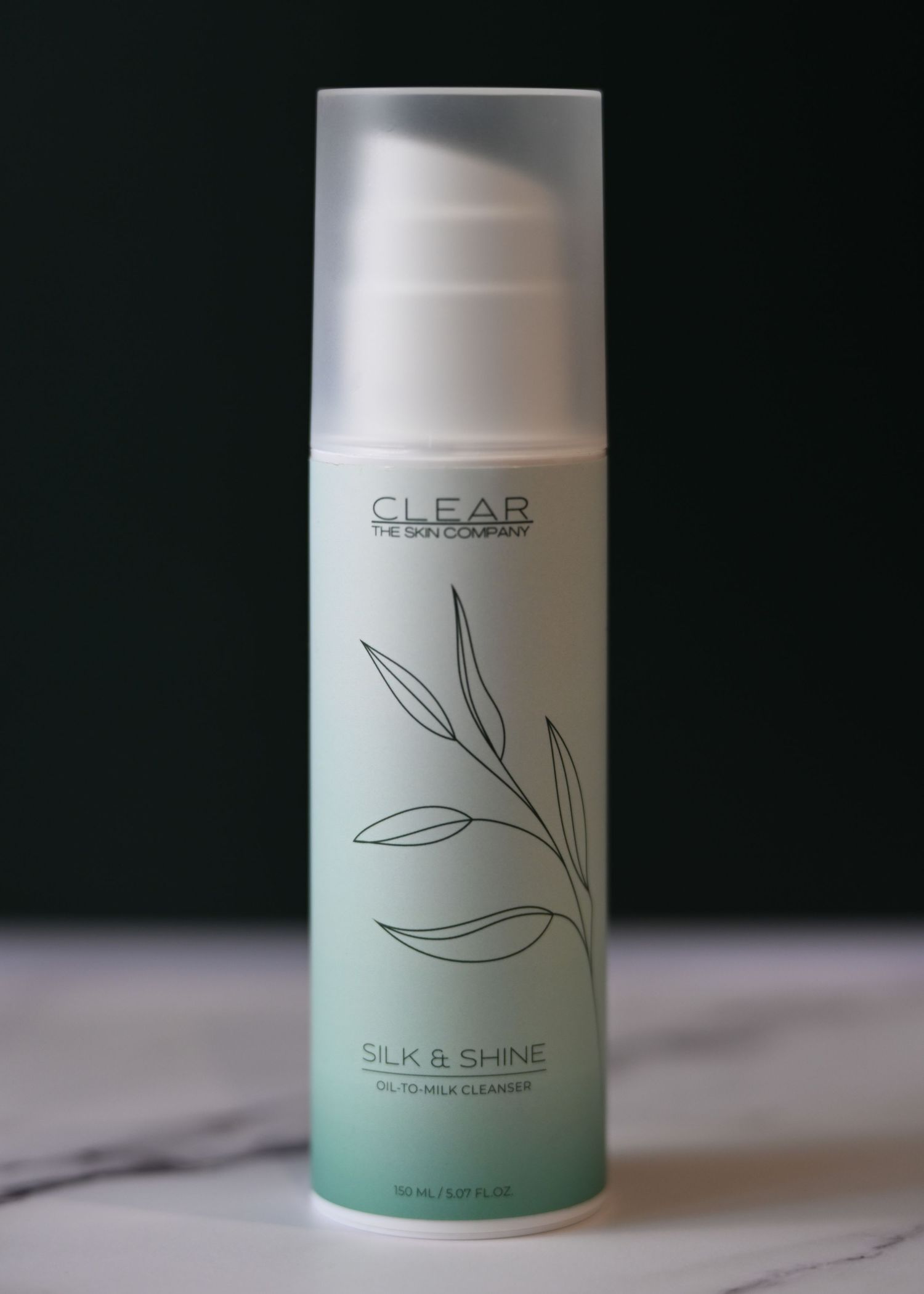 Clear The Skin Company Silk and Shine oil-to-milk cleanser pump bottle on a marble surface with dark background.