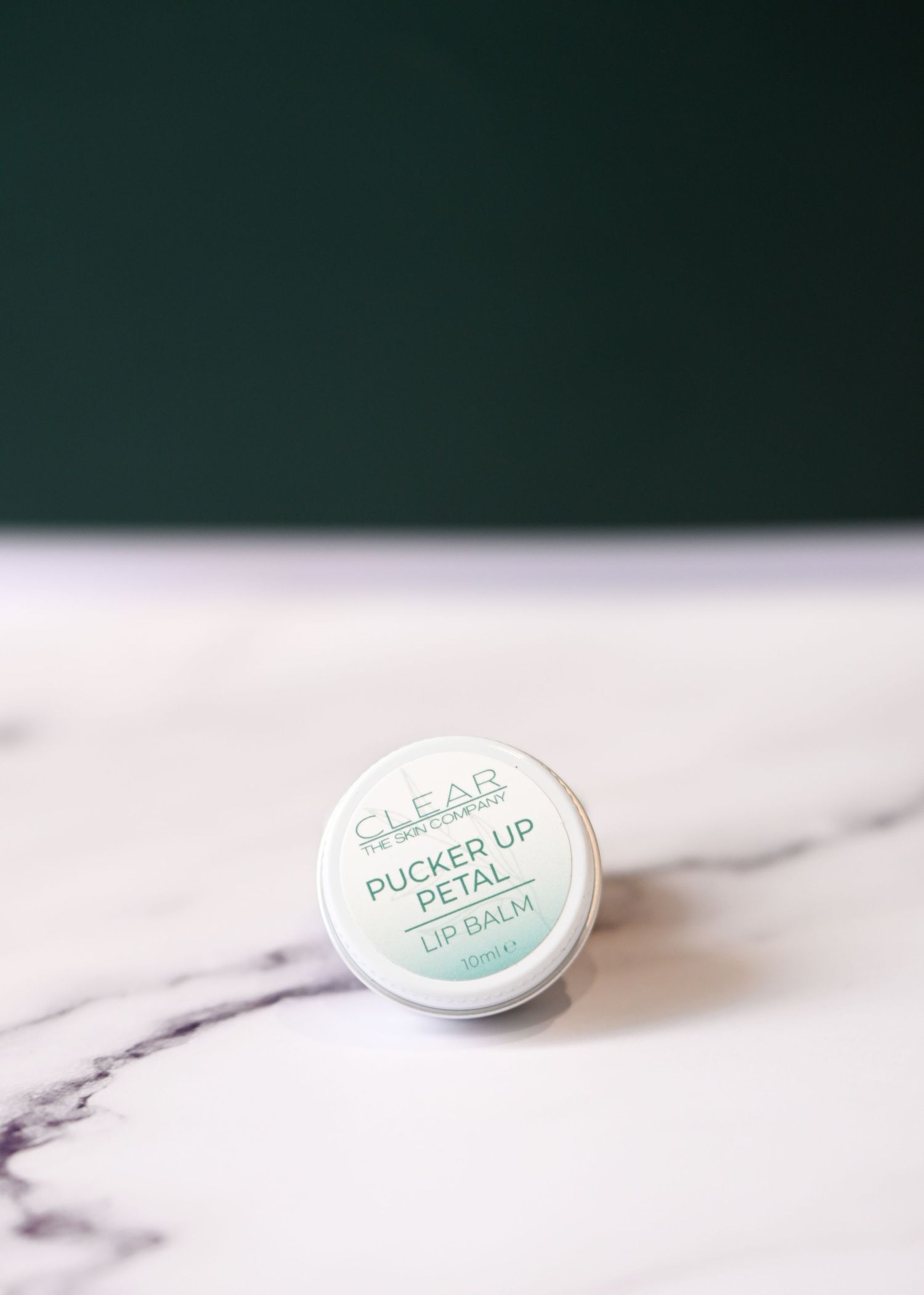 CLEAR Pucker Up Petal lip balm tin with mint green gradient label on white marble surface.