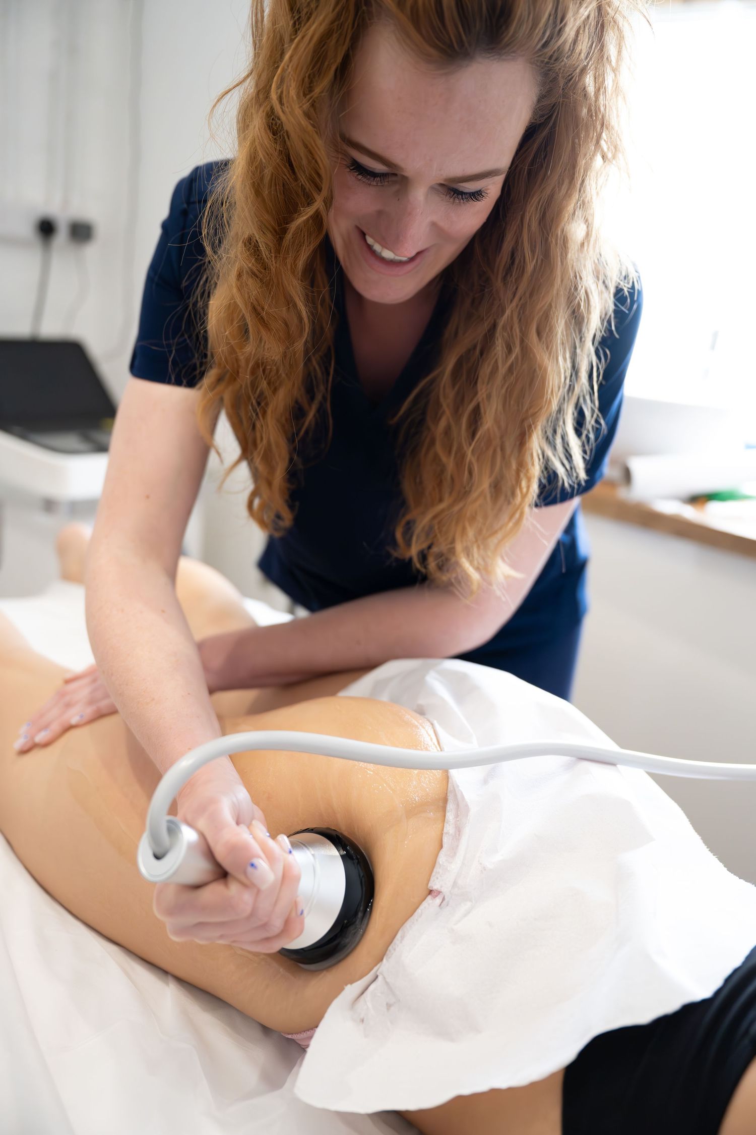 Smiling female aesthetician performing radiofrequency body contouring treatment on client in clinic.