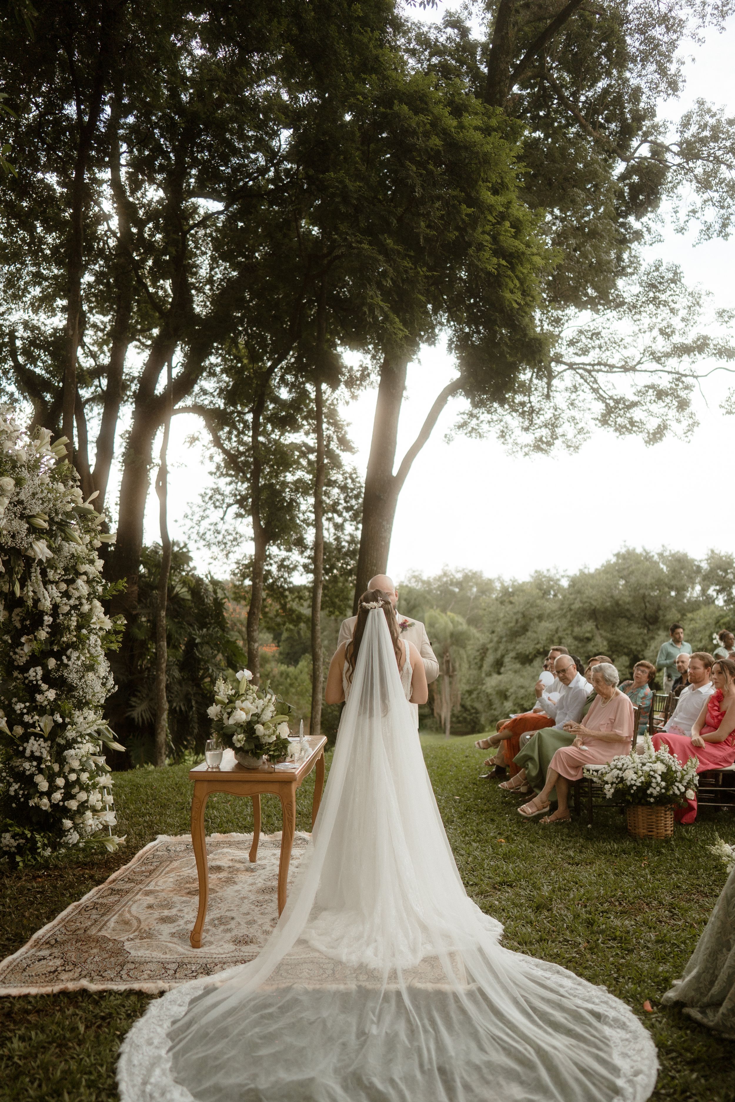 Wedding ceremony scene with flowing white dress train spread across green lawn.