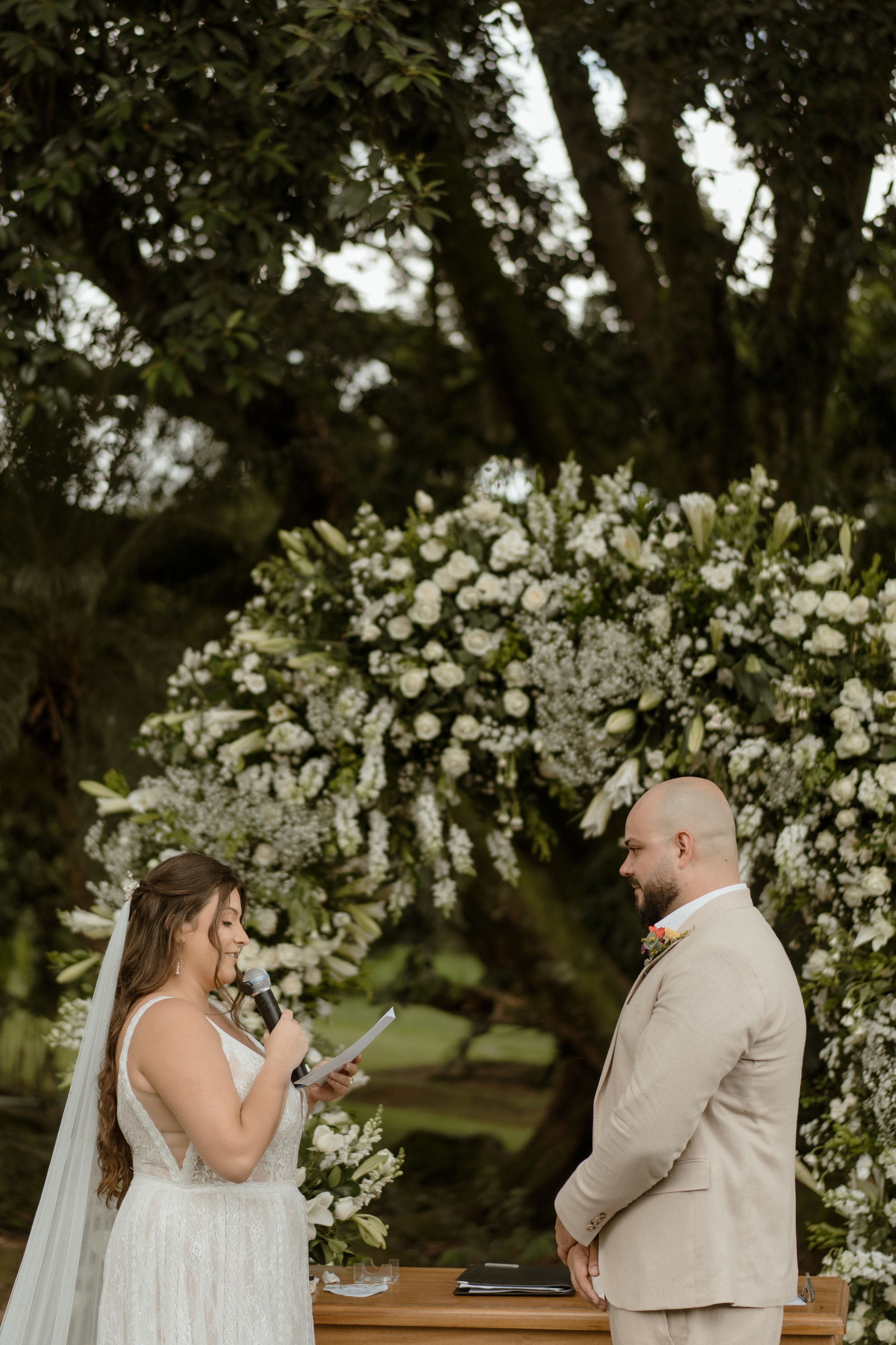 Wedding ceremony under trees with white floral arch and emotional exchange.