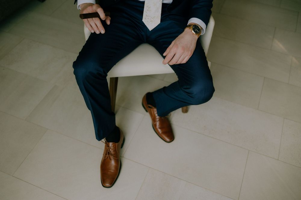 A person in a navy suit and brown leather shoes sitting on a light tile floor with one leg crossed.
