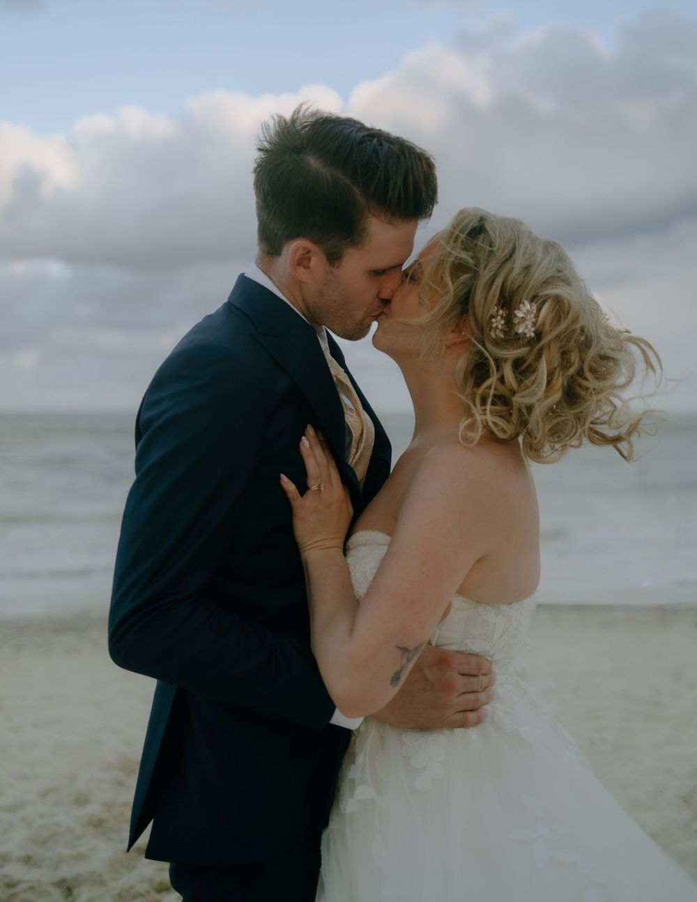 A romantic beach wedding photo shows a couple sharing a tender kiss against a cloudy oceanfront backdrop.