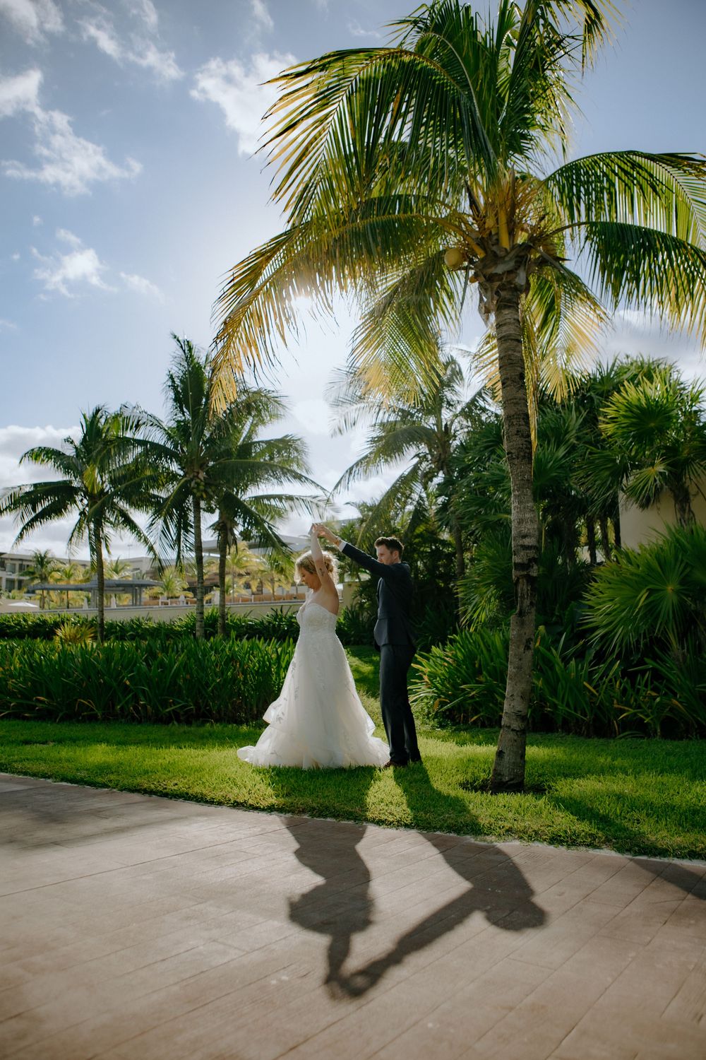 A tropical beach wedding couple stands beneath swaying palm trees casting dramatic shadows on a tiled pathway.