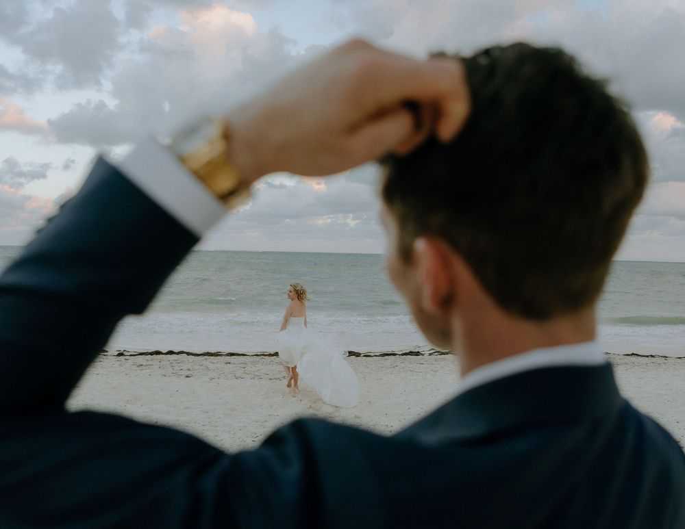 Someone in a suit watches a distant figure in white walking along a beach at sunset.