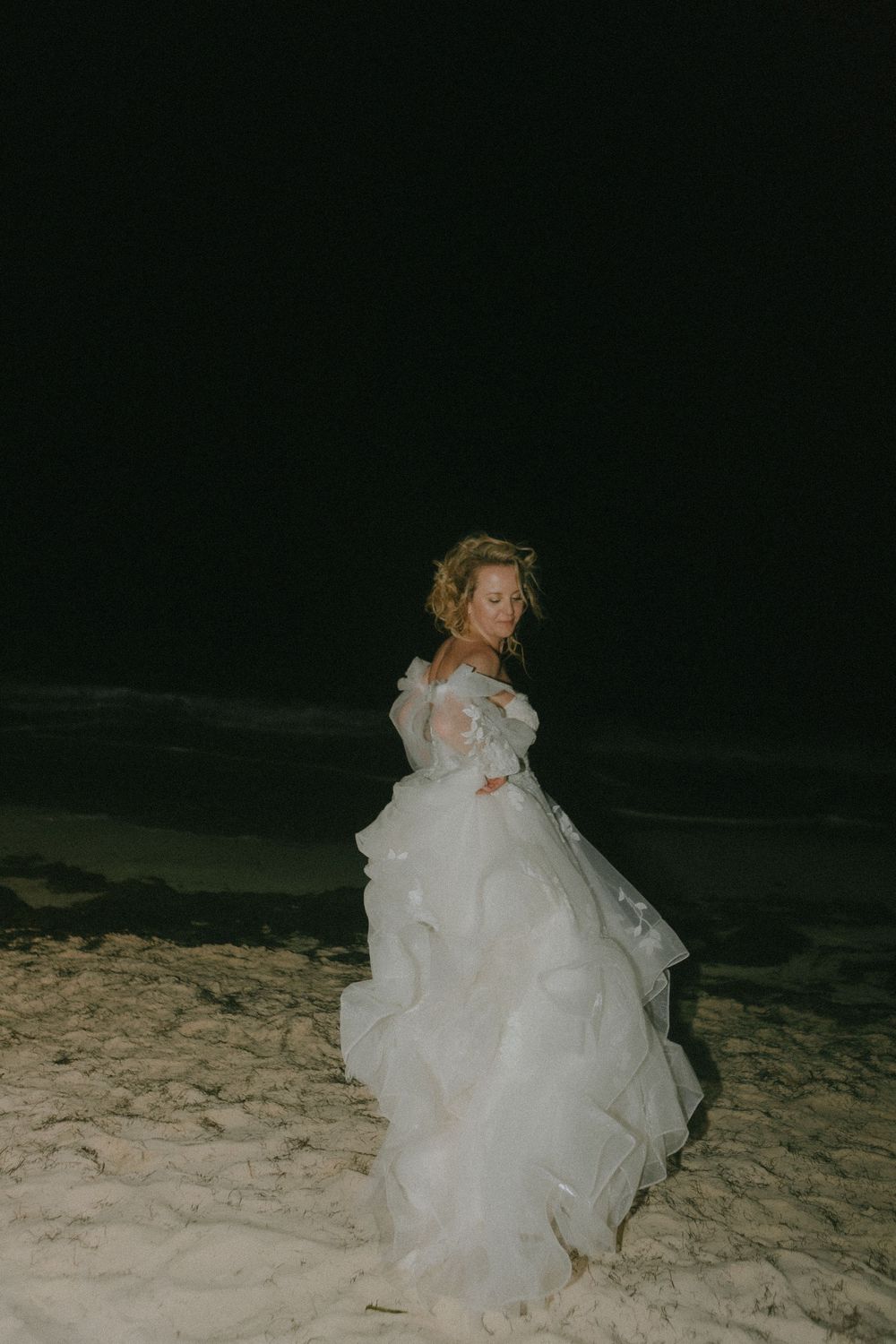 A bride in a flowing white gown poses on a sandy beach at night.
