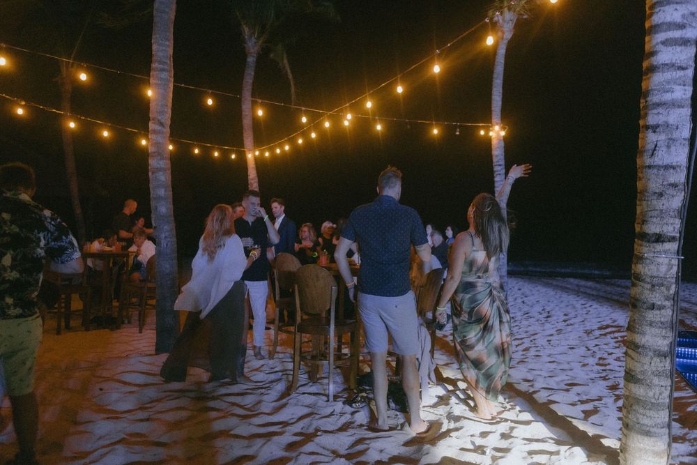People gathered for a nighttime beach party with string lights illuminating the sandy shore and palm trees.