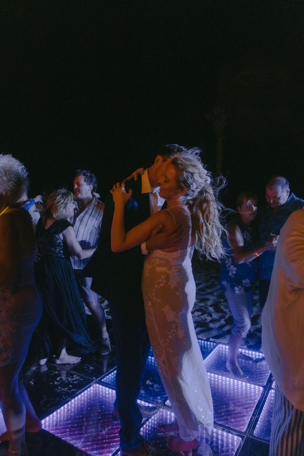 Wedding guests dance on an illuminated dance floor during an evening reception.