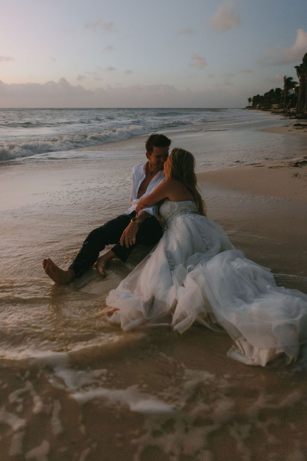 A romantic couple sitting on a beach at sunset with waves lapping at their feet while wearing wedding attire.