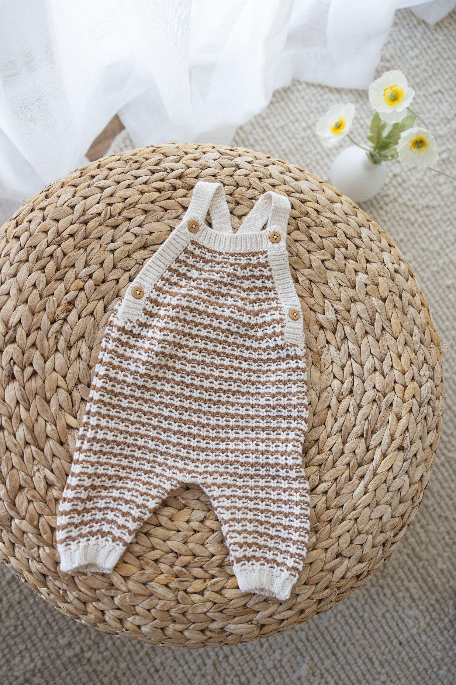 White and tan striped knitted baby overalls displayed on a round woven rattan mat with small white flowers in background.