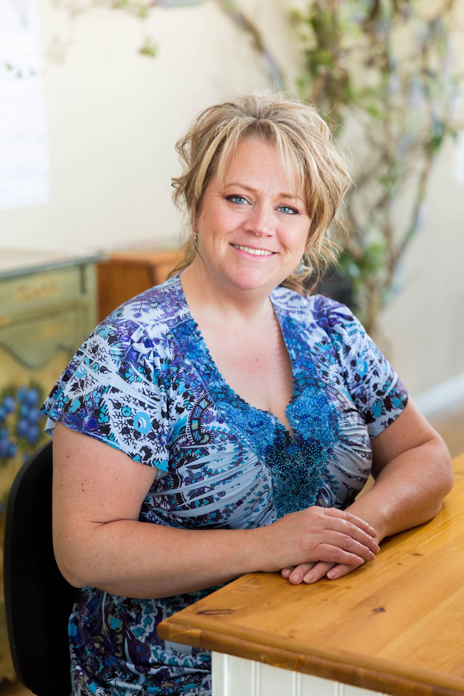 A woman in a blue patterned top sits at a wooden table with a warm smile.