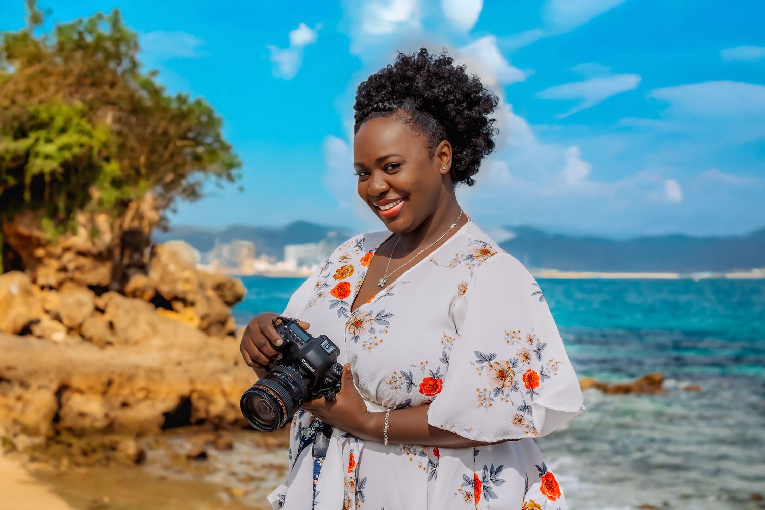 A photographer in a floral dress reviews images on their camera near a tropical coastline.