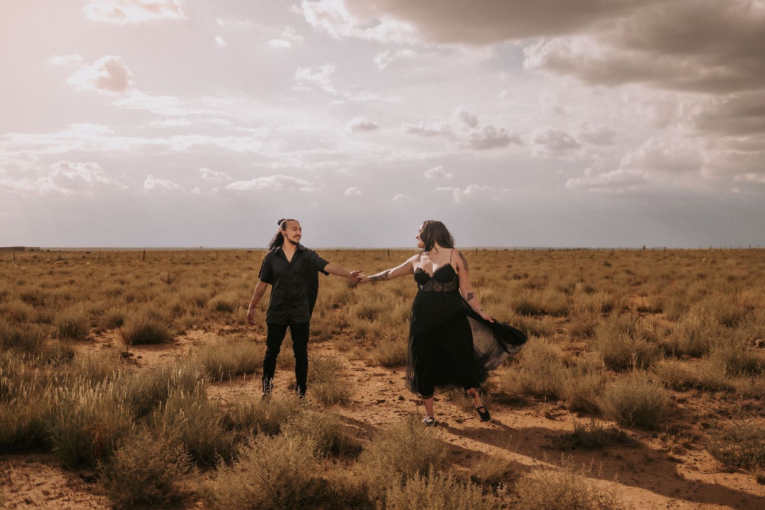 a queer couple dances and spins together in an open field under dramatic cloudy skies at sunset in the desert in Albuquerque, New Mexico.