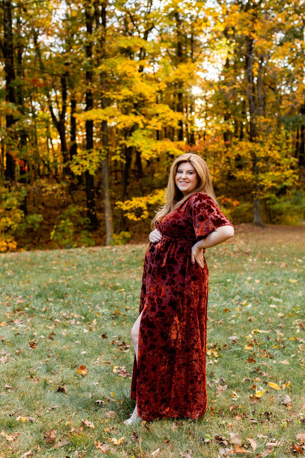 A pregnant person wearing a burgundy velvet dress poses in an autumn forest with golden leaves in the background. Maternity NJ photographer