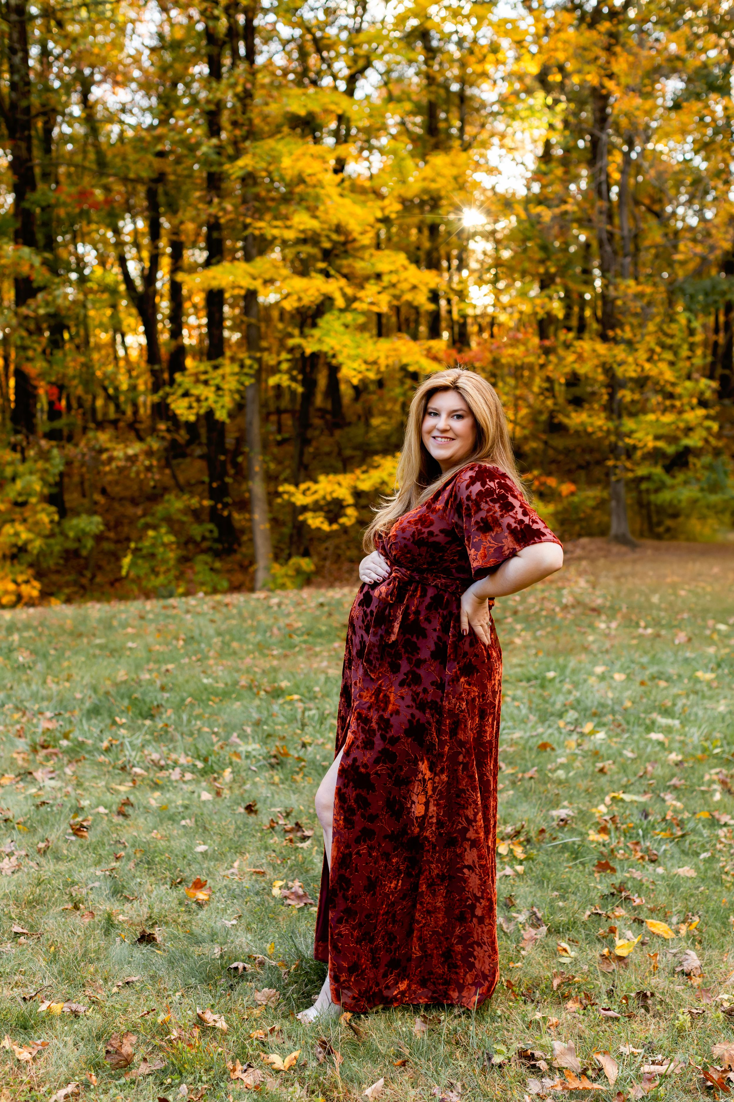 A pregnant person wearing a burgundy velvet dress poses in an autumn forest with golden leaves in the background. Maternity NJ photographer