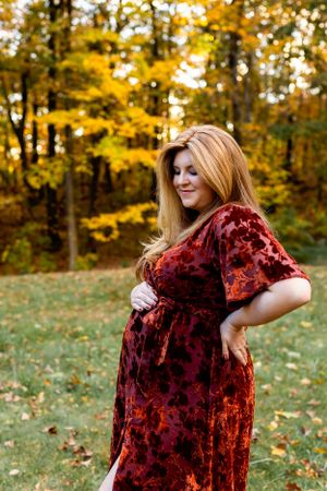 A person in a burgundy velvet dress poses outdoors against a backdrop of golden autumn foliage. Maternity NJ photographer