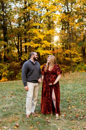 A couple stands together in a fall forest with vibrant yellow foliage and sunlight peeking through the trees. Maternity NJ photographer