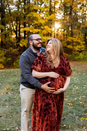 A maternity photo shows a couple embracing outdoors in autumn, with the mother wearing a burgundy velvet dress. Maternity NJ photographer