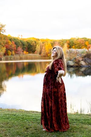 A velvet burgundy maternity dress is worn by someone posing near an autumn lake with colorful fall foliage. Maternity NJ photographer