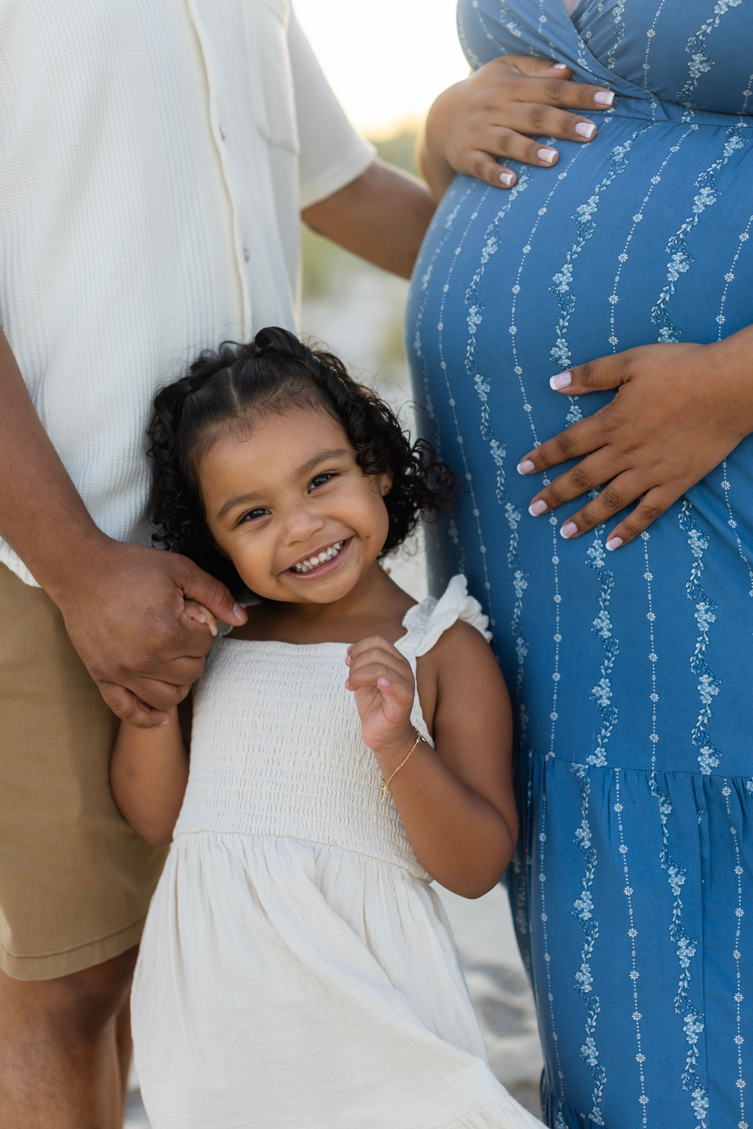 A smiling toddler girl in white dress stands between her parents, with pregnant mother in blue floral dress holding her baby bump.