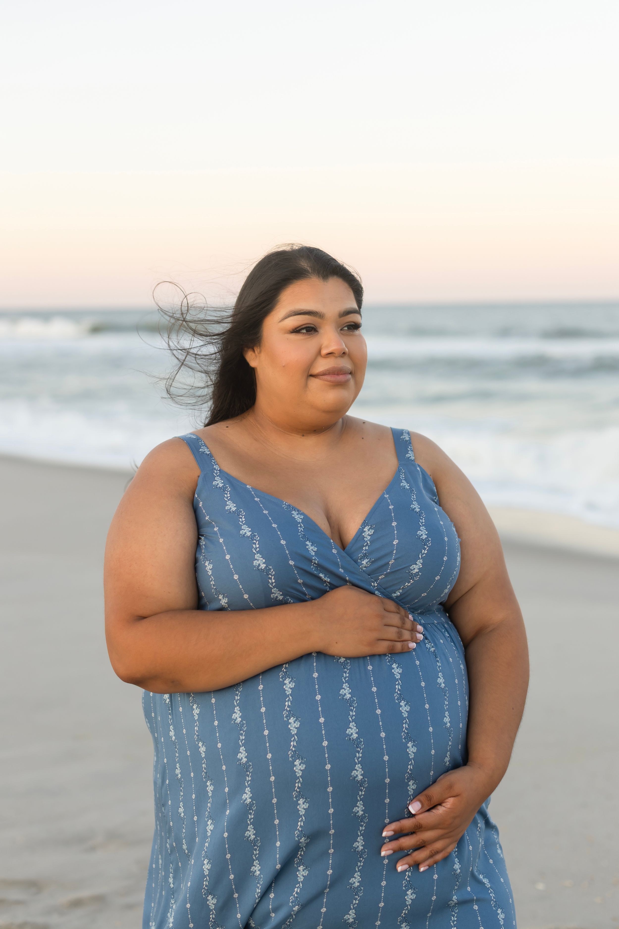 Plus-size woman in blue floral sundress smiling at beach during sunset, wind blowing her dark hair.