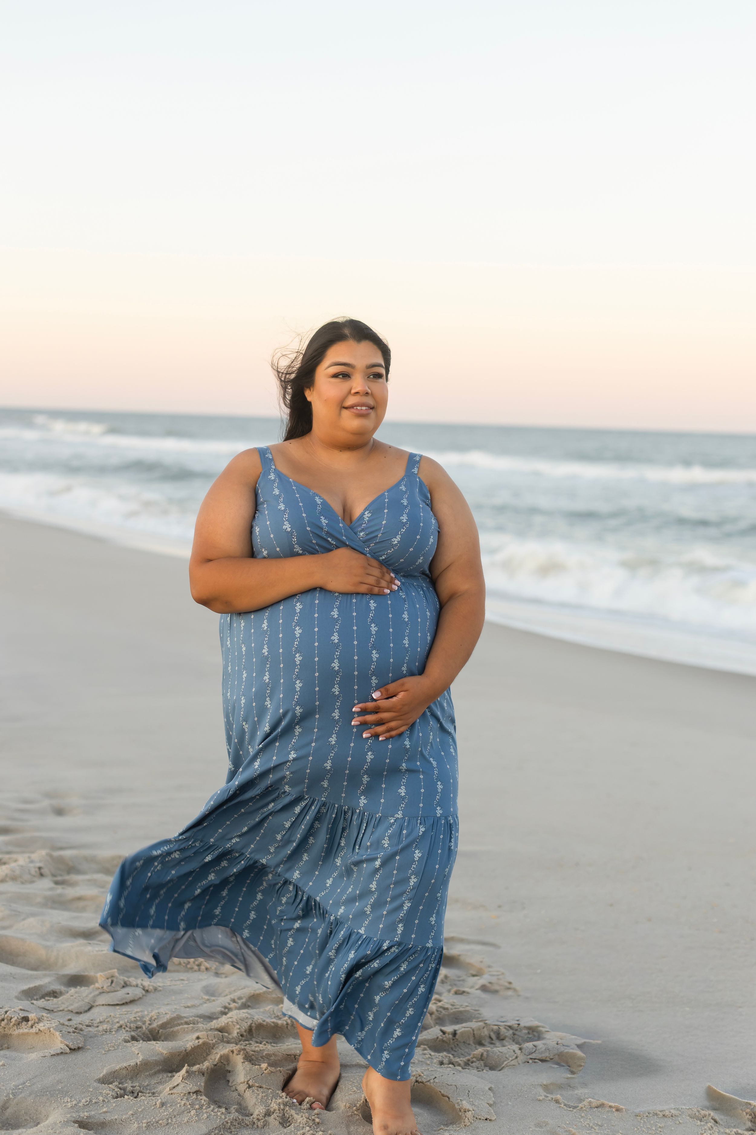 A plus-size woman in a blue floral maxi dress walks barefoot on a sandy beach at sunset.