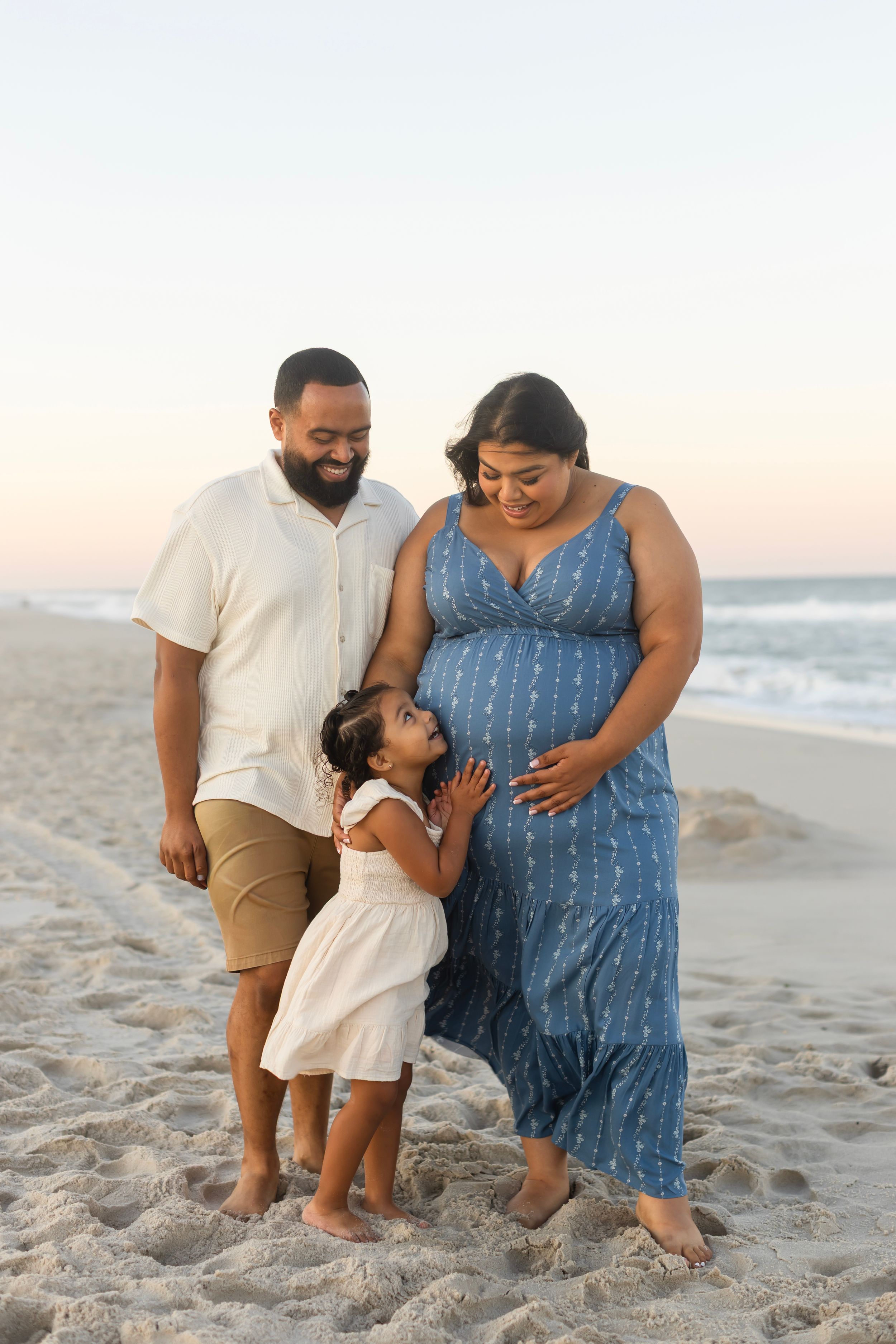 Pregnant woman in blue dress with husband and toddler daughter enjoying a family beach photoshoot at sunset.