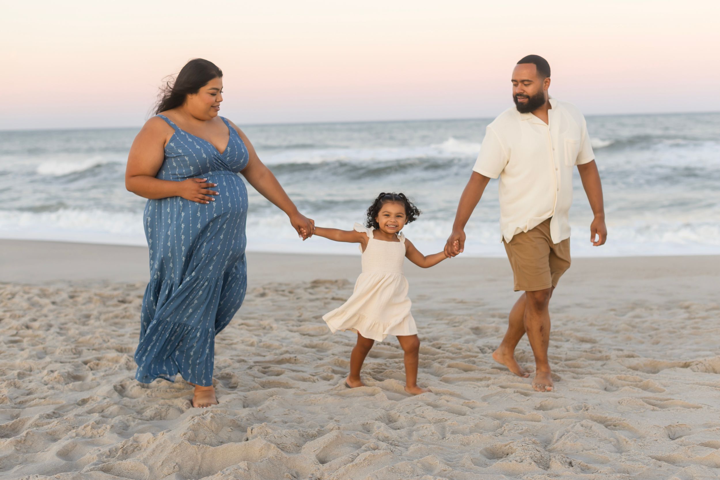 Family of three walking on beach at sunset, parents holding young daughter's hands near ocean waves.