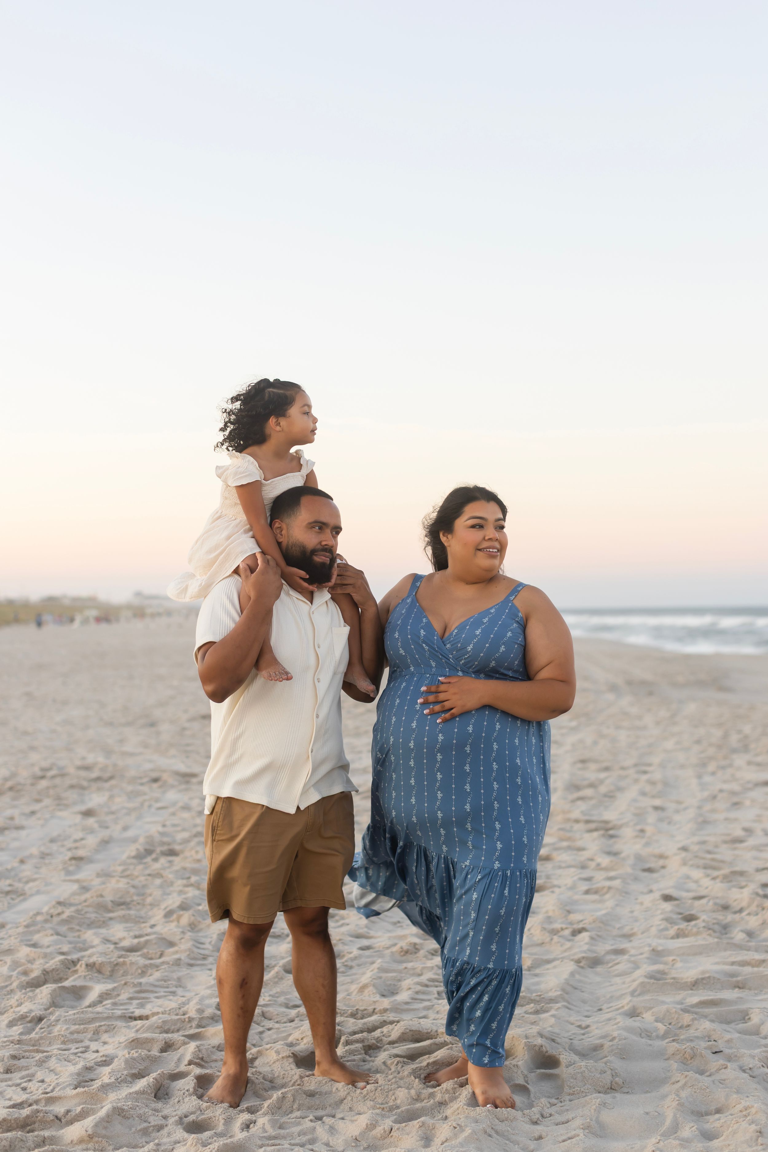 Family of three enjoying a sunset beach walk, with child on father's shoulders beside pregnant mother in blue dress.