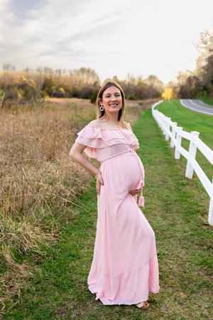 Soon to be mama in a flowing maternity dress stands along a white fence and grassy path at sunset. Sussex County Maternity NJ Photographer