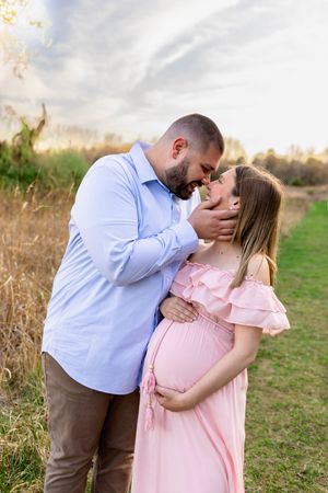 Soon to be mama in pink dress embraces partner in blue shirt on grassy field at sunset. NJ Maternity Photographer