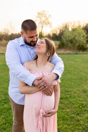 A couple embraces in a grassy field during golden hour while wearing a pink dress and blue button-down shirt. NJ Maternity Photographer