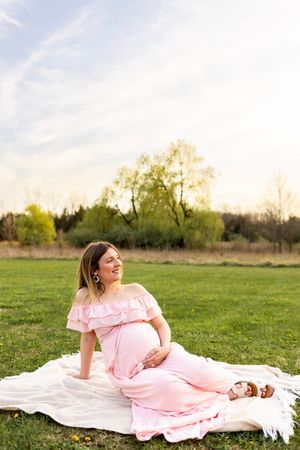 A person in a pink off-shoulder maternity dress relaxes on a white blanket in a sunny field at sunset. NJ Maternity Photographer