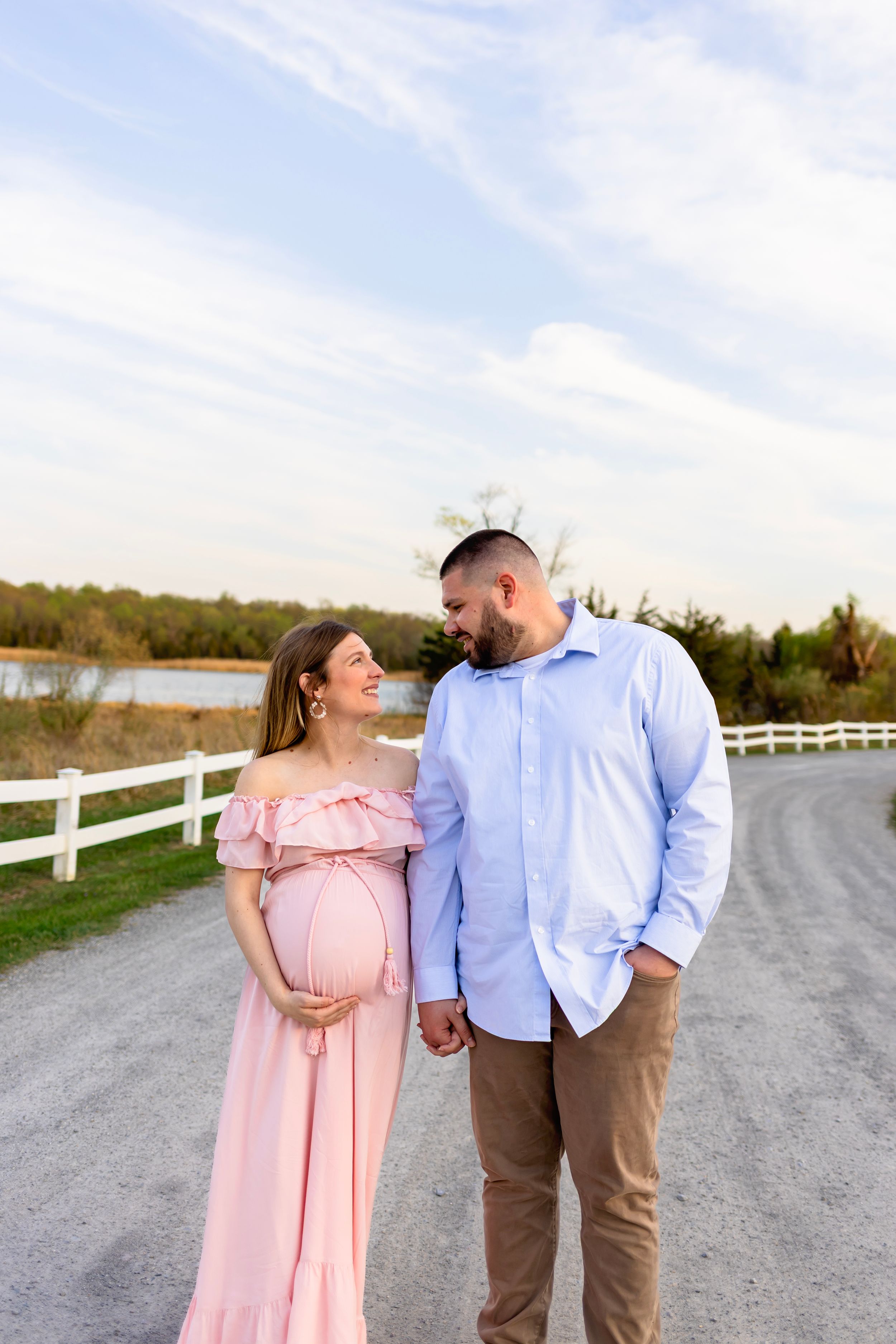 A couple shares a romantic moment on a country path with white fencing and a lake in the background. NJ Maternity Photographer
