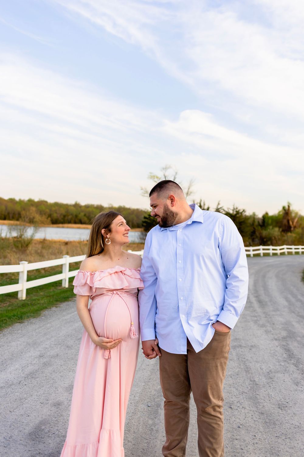 A couple shares a romantic moment on a country path with white fencing and a lake in the background. NJ Maternity Photographer