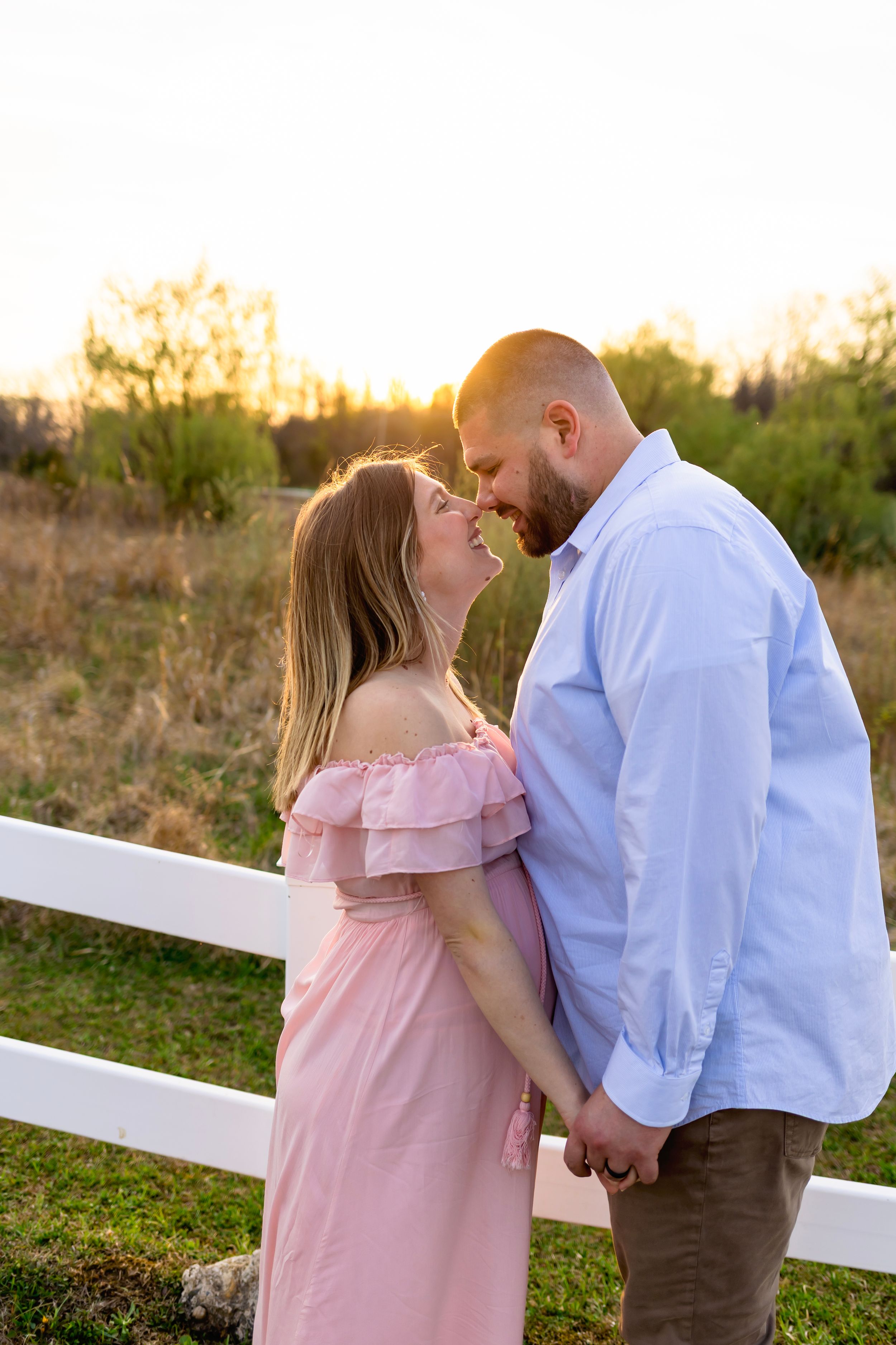 NJ Maternity Photographer couple shares an intimate moment at sunset by a white fence in a pastoral setting with pink dress and blue shirt.