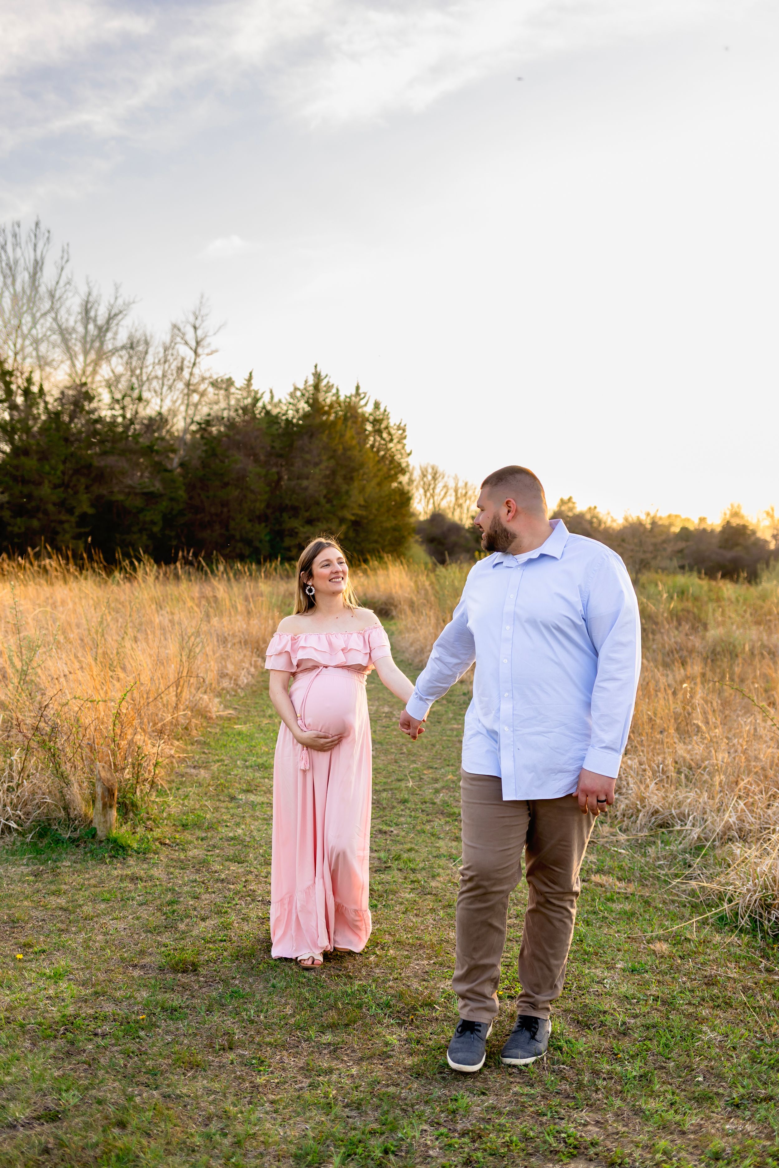 A couple stands together in a golden field at sunset, with the woman wearing a flowing pink dress. NJ Maternity Photographer