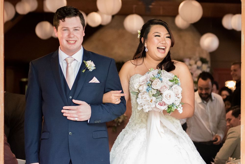A joyful wedding couple shares a laugh while walking down the aisle with string lights in background.
