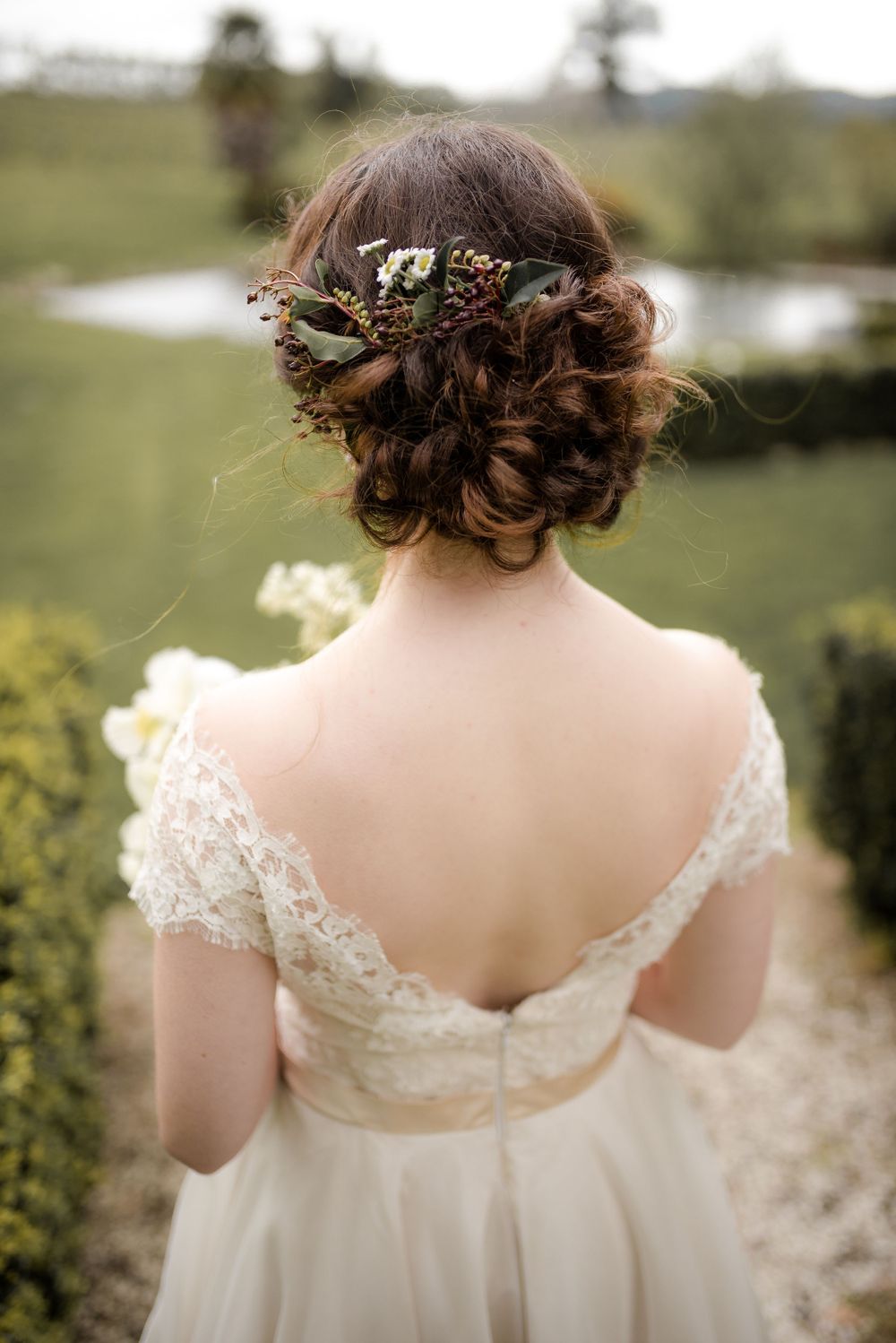 Back view of a wedding dress showing intricate lace detail and floral hair accessories against a garden backdrop.