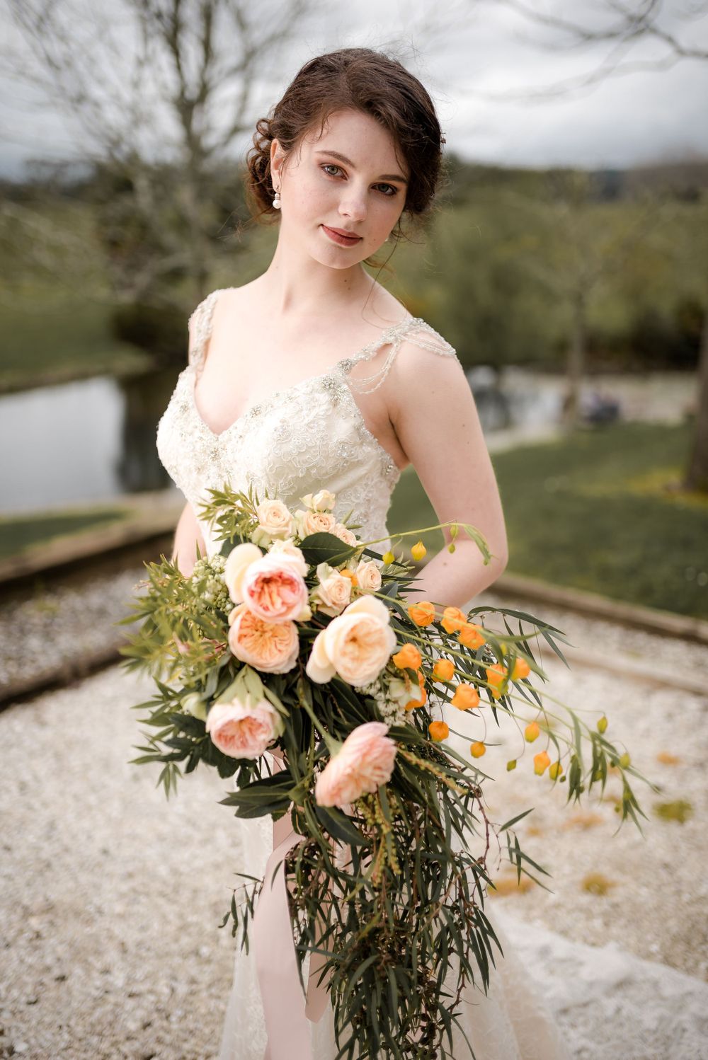 Romantic bridal portrait showcasing a delicate lace gown and fresh peach-colored floral bouquet in an outdoor setting.