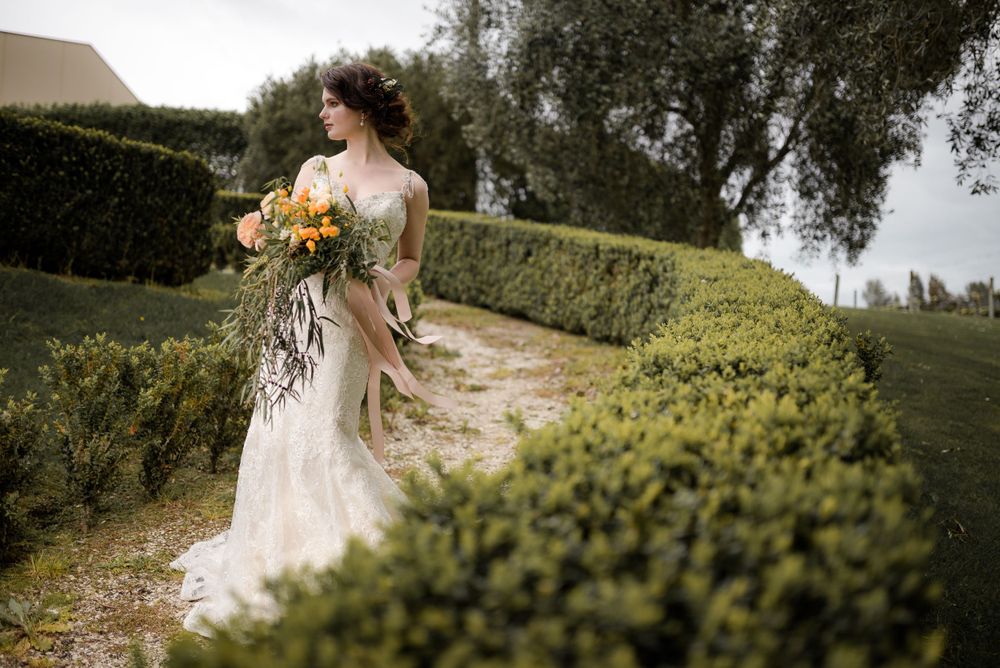A bride in a lace wedding dress walks through a garden path holding a cascading bouquet of peach and orange flowers.