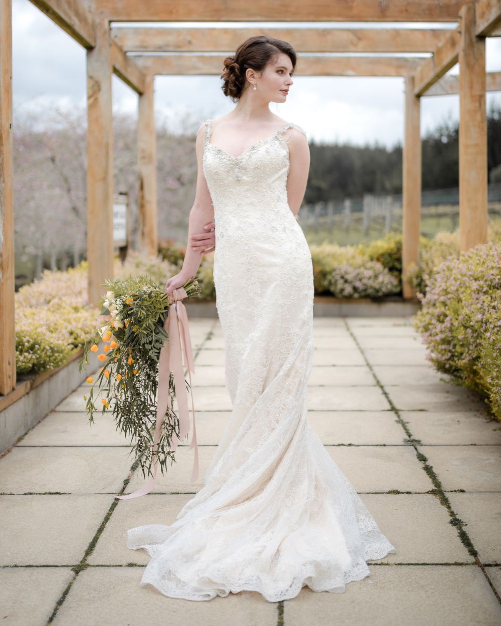 A bride in a fitted lace wedding gown poses under a wooden pergola with cascading bouquet.