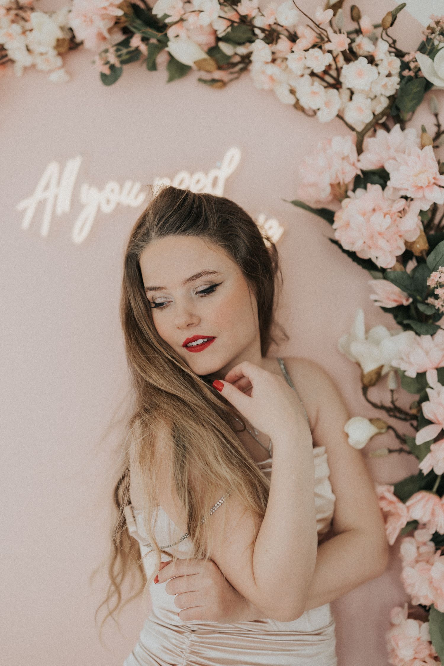 Beautiful woman in white dress posing under floral arch with All You Need neon sign.