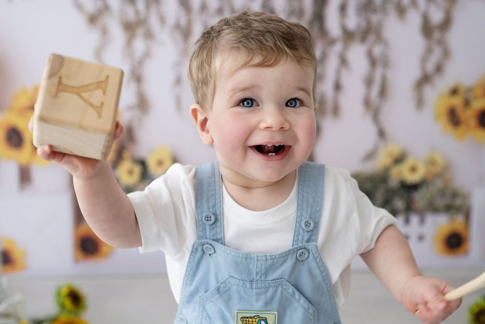 Happy toddler in light blue overalls holds up a wooden block while playing with sunflower decorations in background.