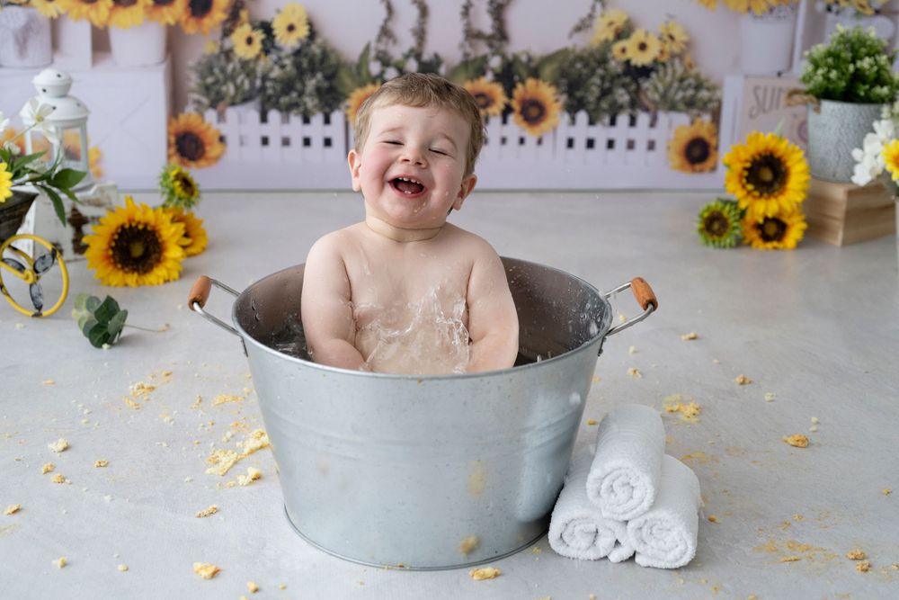 A happy baby sits in a metal wash basin surrounded by bright yellow sunflowers.