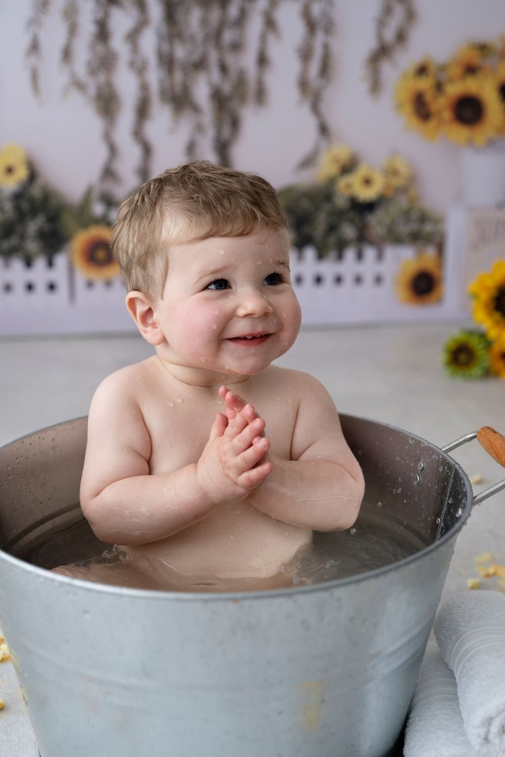 Cake Smash Photography Melbourne, a happy baby sits in a metal wash tub for a bath with sunflower decorations in the background.