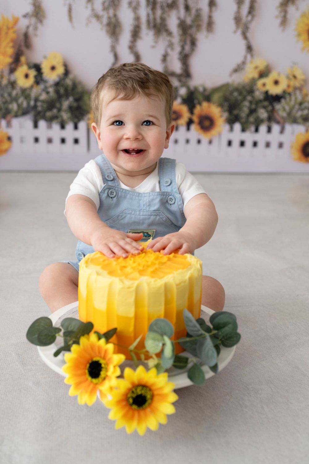 Cake Smash Photography, toddler sitting behind a yellow birthday cake with sunflower decorations on white floor with floral backdrop.