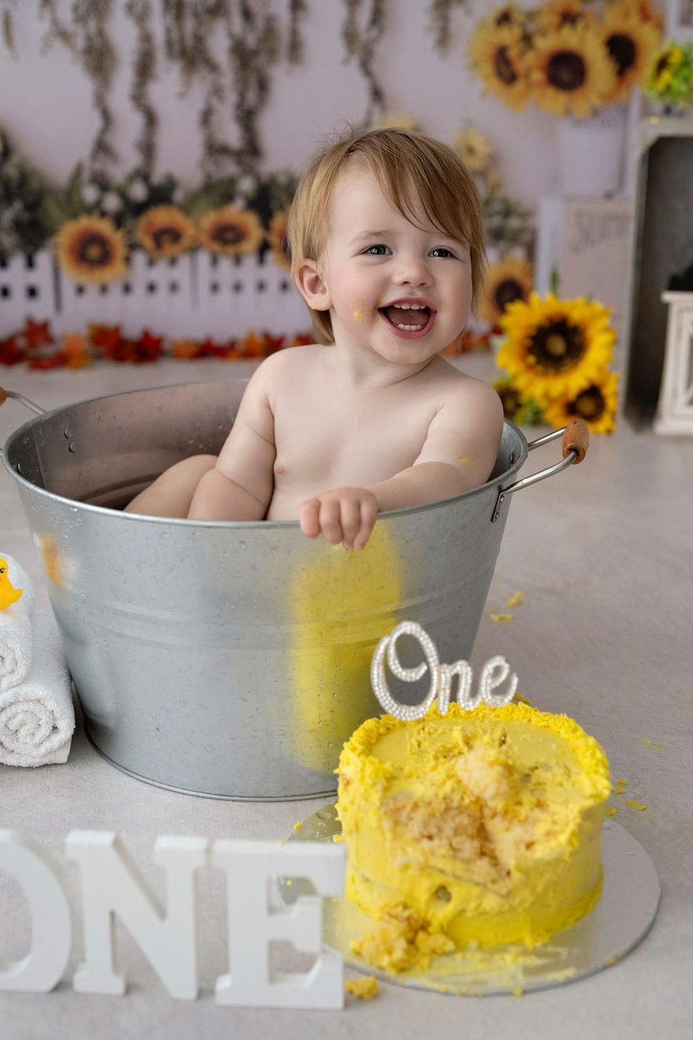 Happy, smiling baby sitting in metal tub with yellow cake smash and sunflower decorations for first birthday photoshoot.