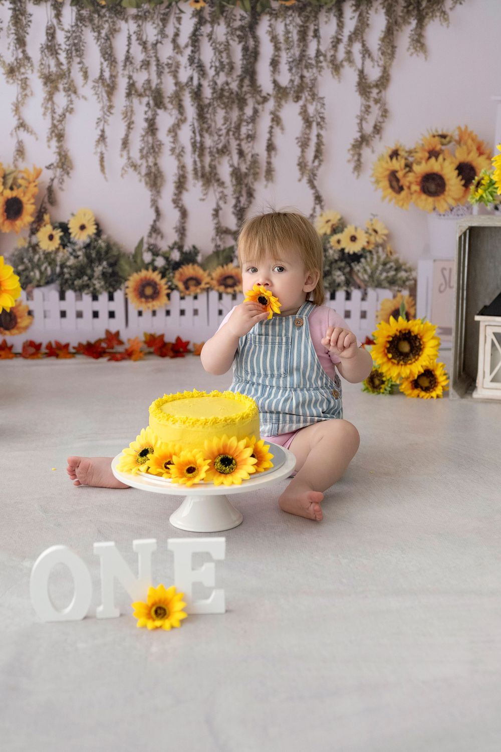 A cake smash photo session featuring bright sunflowers and a white cake against a rustic backdrop.