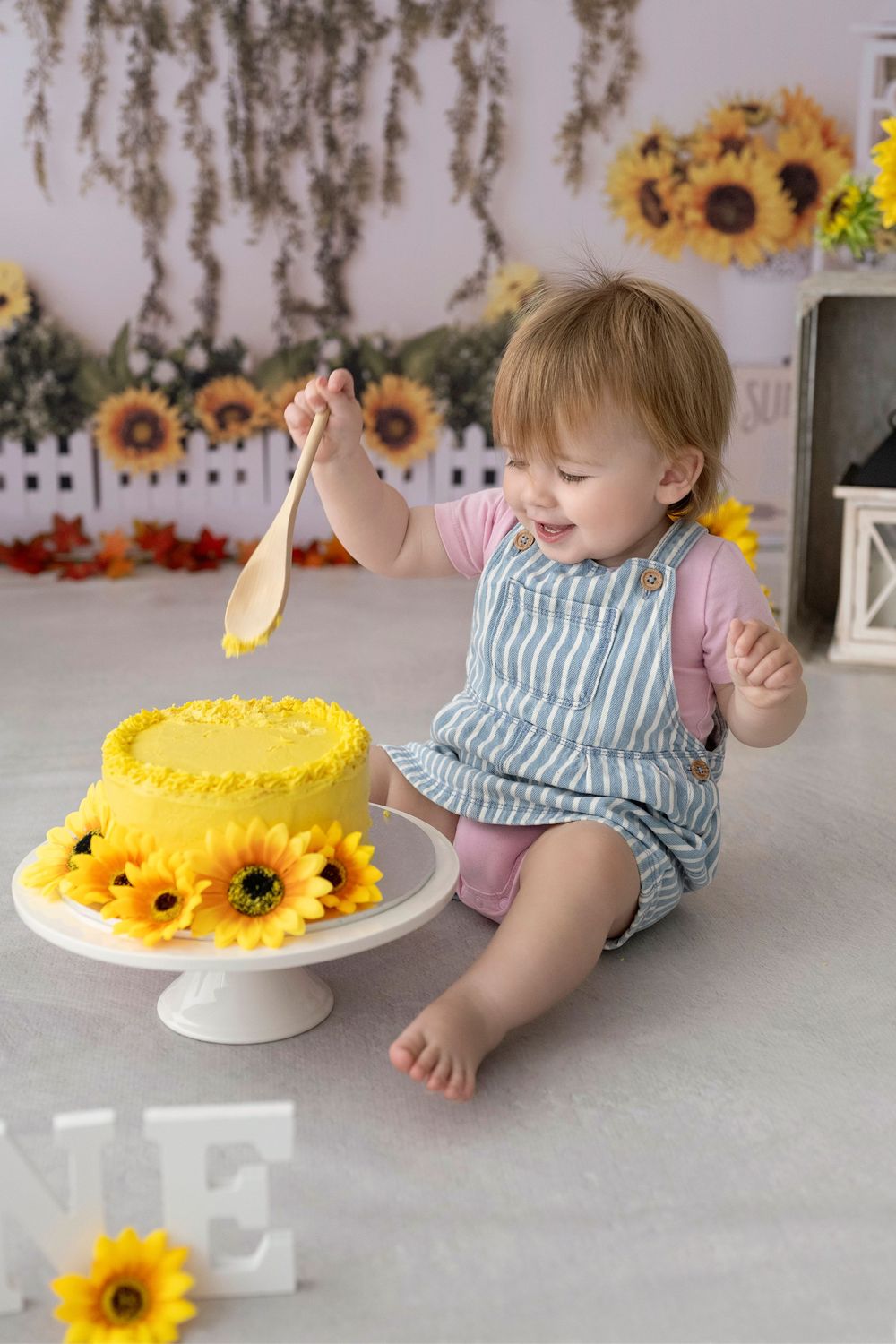 Baby in striped outfit sits near yellow birthday cake decorated with bright sunflowers in a bright studio setting.